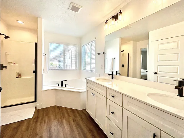 Bathroom featuring wood-finish flooring, a double vanity with white cabinetry and integrated sinks, a built-in corner tub with black fixtures, and a glass-enclosed shower with black hardware