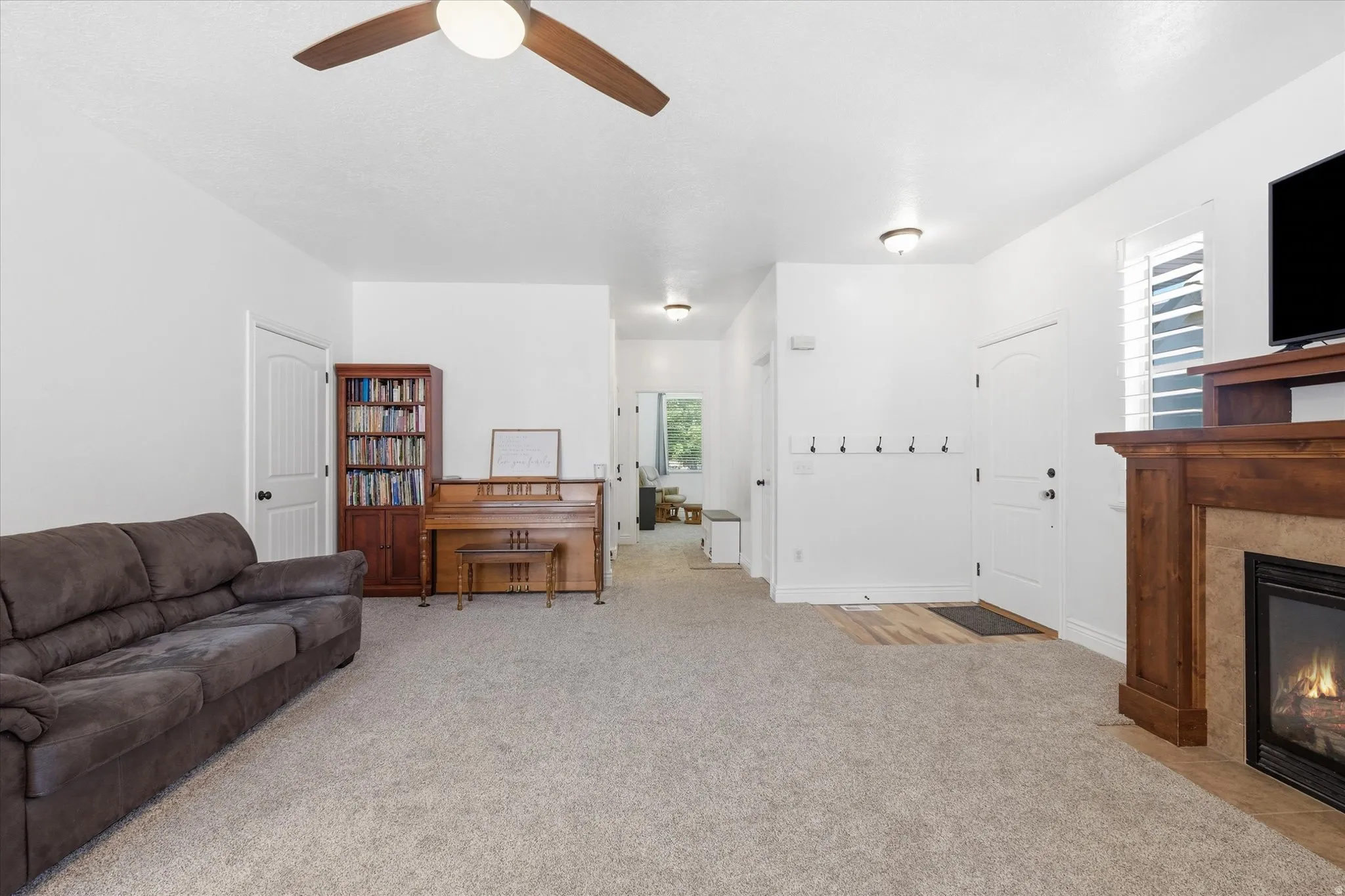 Carpeted living area featuring a fireplace with a wood-finish mantel and stone surround, a ceiling fan, and a front entry door with a coat rack