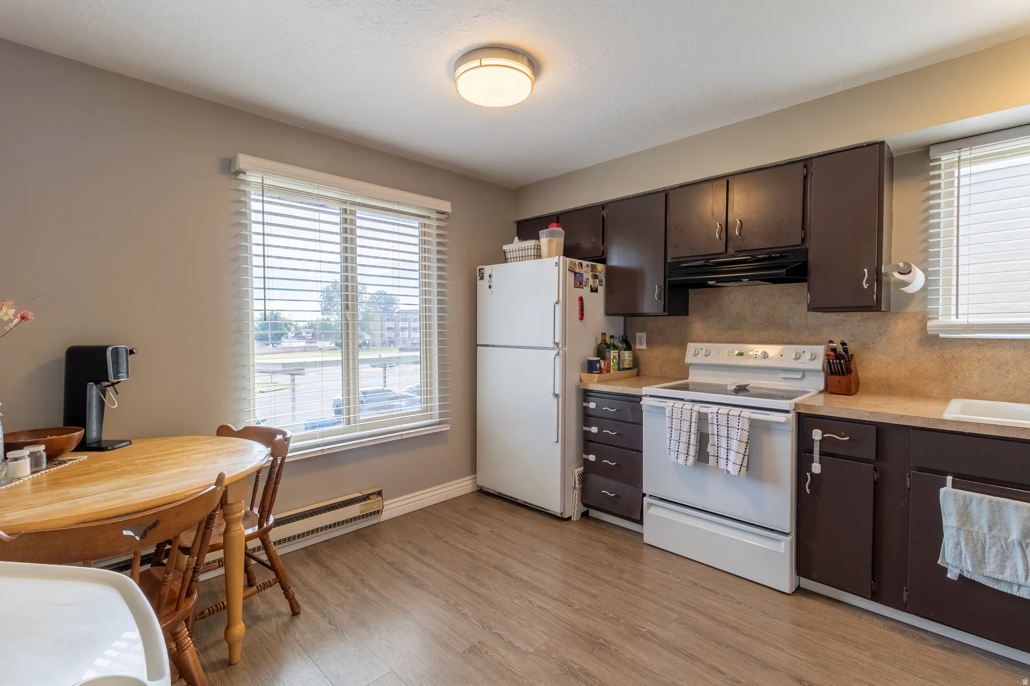 Kitchen featuring wood-finish flooring, dark wood cabinetry, white appliances, and a window with blinds