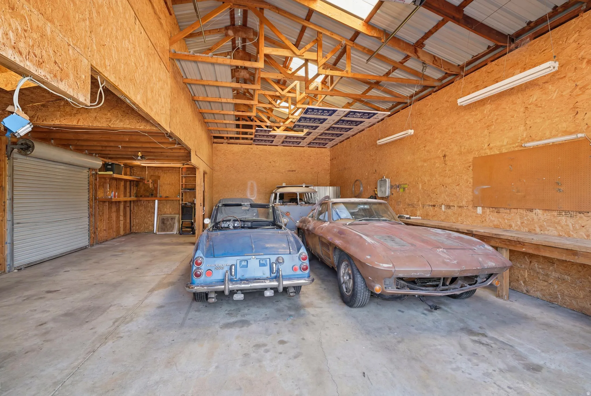 Spacious garage interior featuring concrete flooring, exposed wood beam ceiling, corrugated metal roofing, skylights, and multiple fluorescent light fixtures