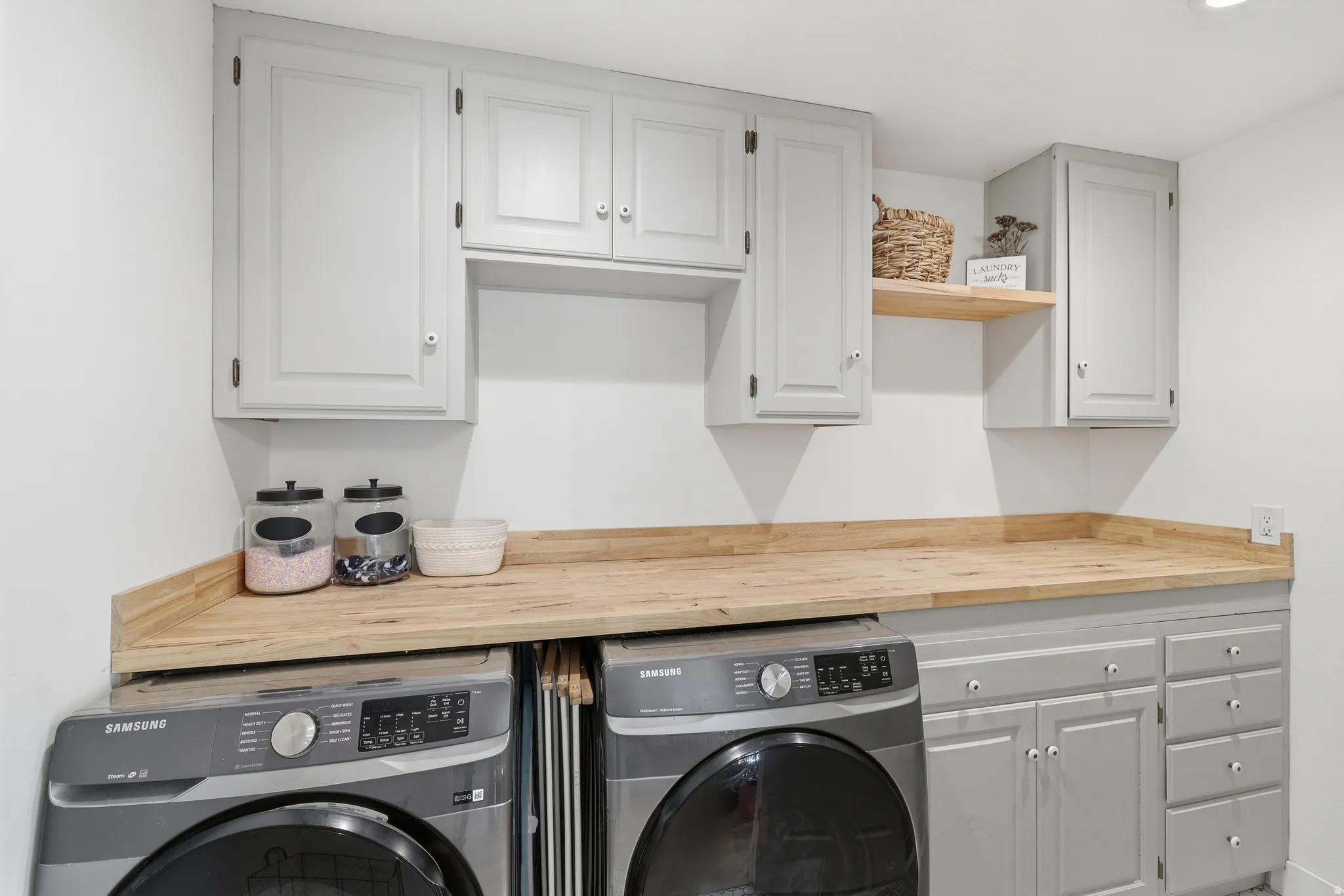 Functional laundry area featuring light gray cabinetry, wood-finish countertops, and an open wood-finish shelf