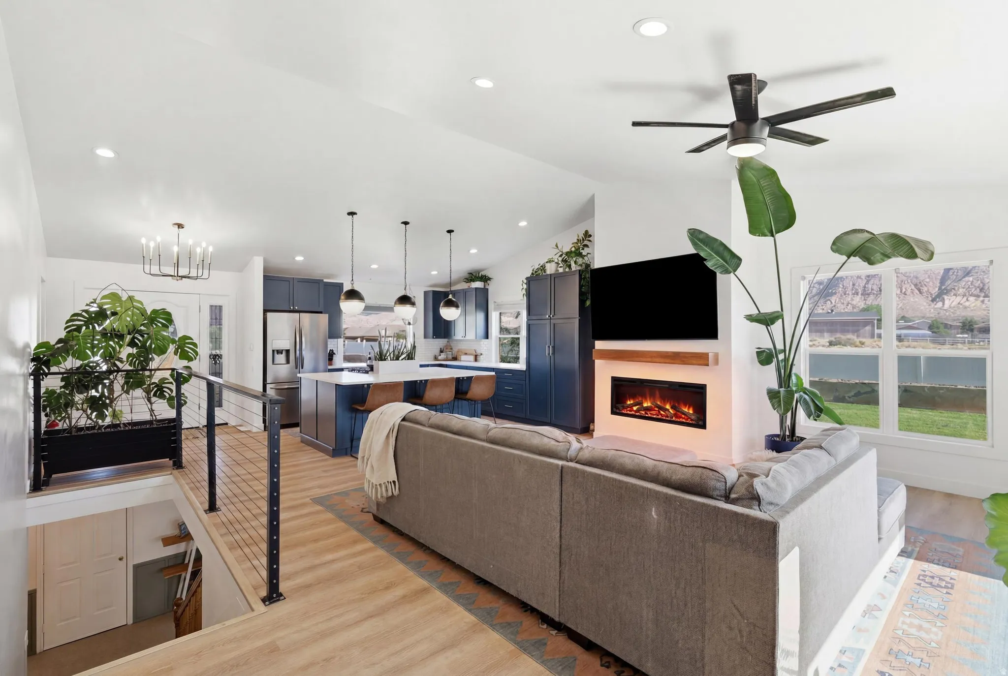 Open-concept living area featuring wood-finish flooring, a contemporary electric fireplace with a wood mantel, a modern ceiling fan, and a kitchen with navy cabinetry and a white island