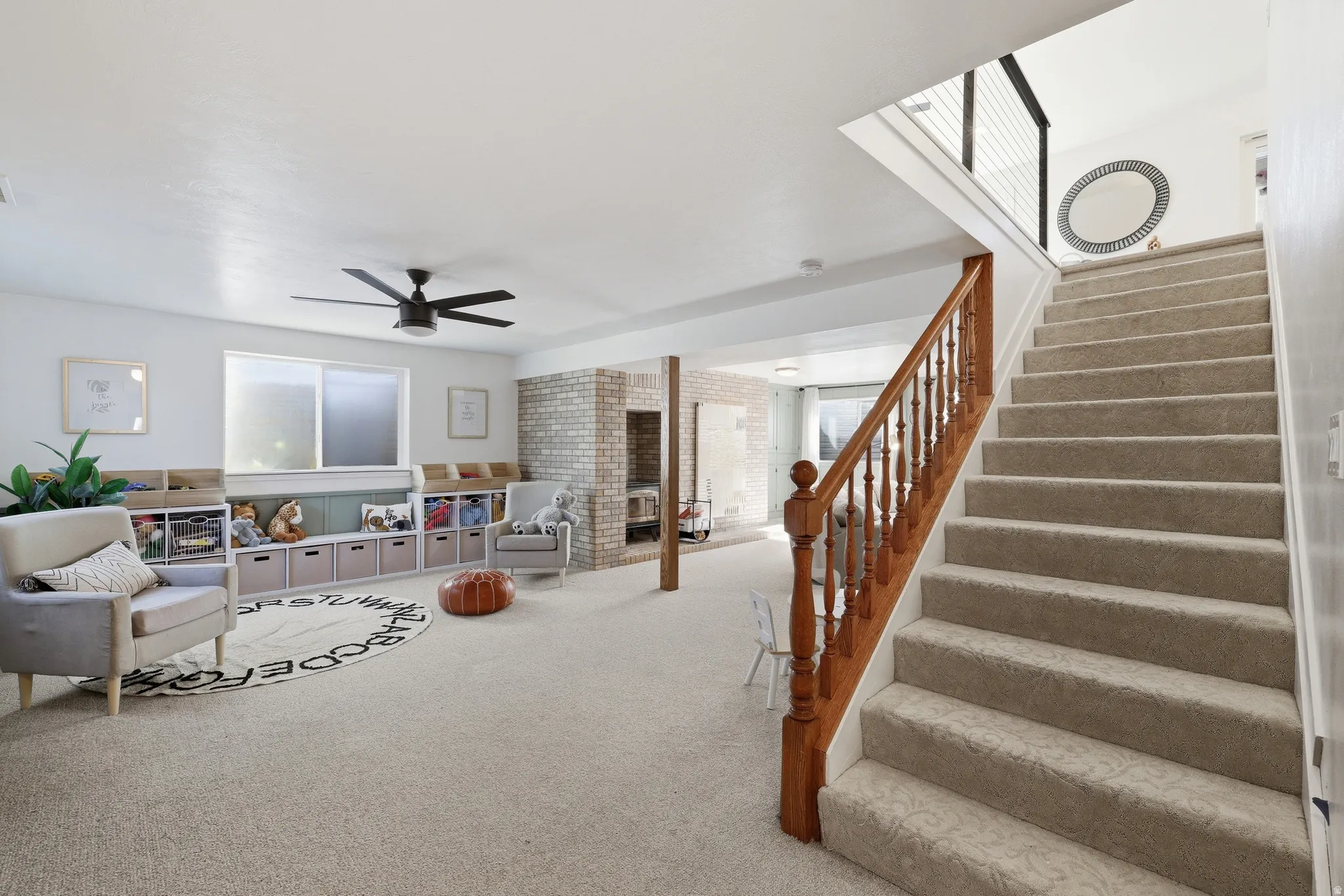 Carpeted staircase with wood newel posts and banister, leading to a lower level featuring recessed lighting, a modern ceiling fan, and a brick accent wall with a built-in fireplace