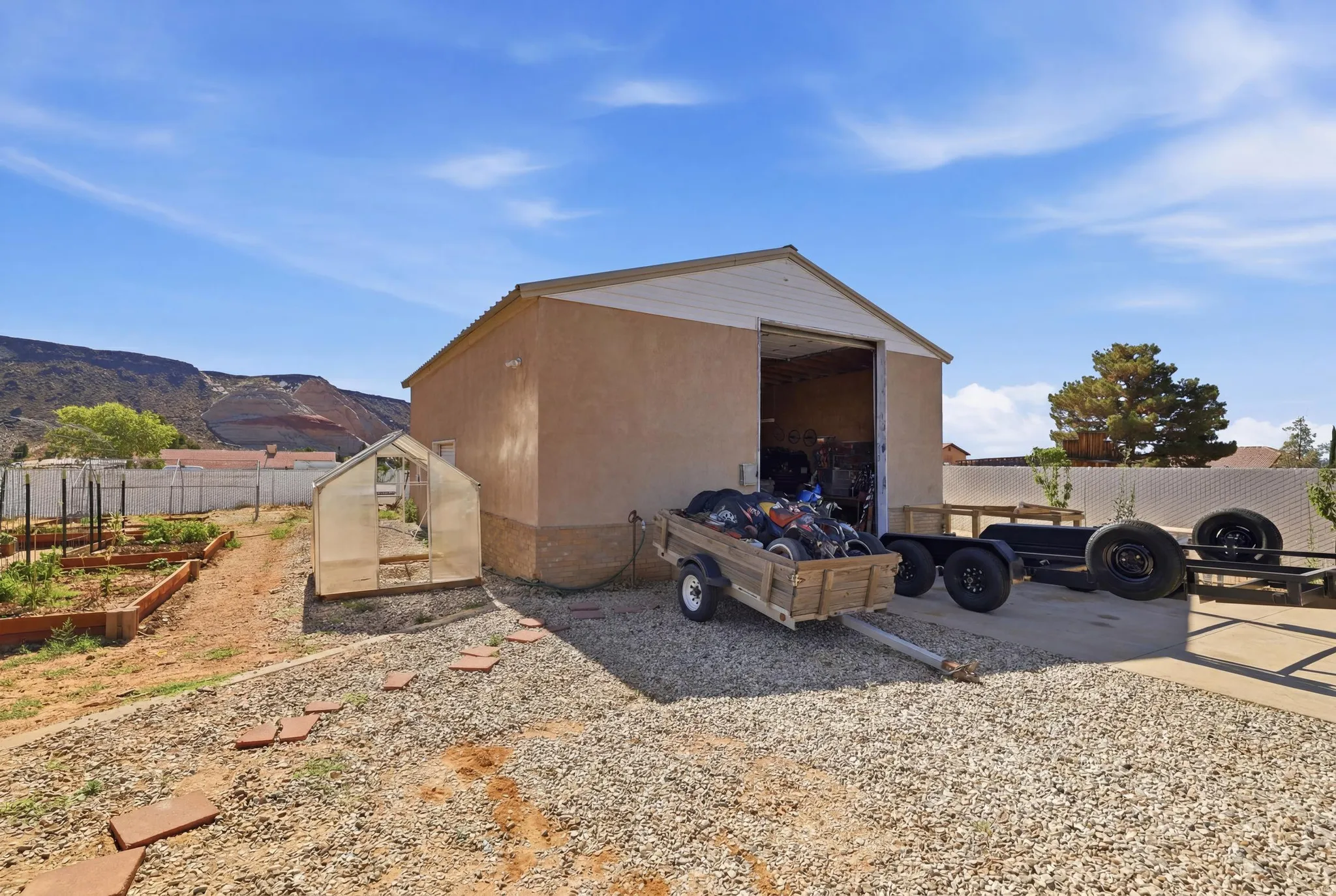 Detached stucco structure with a roll-up door, adjacent to a clear-pane greenhouse and multiple raised garden beds