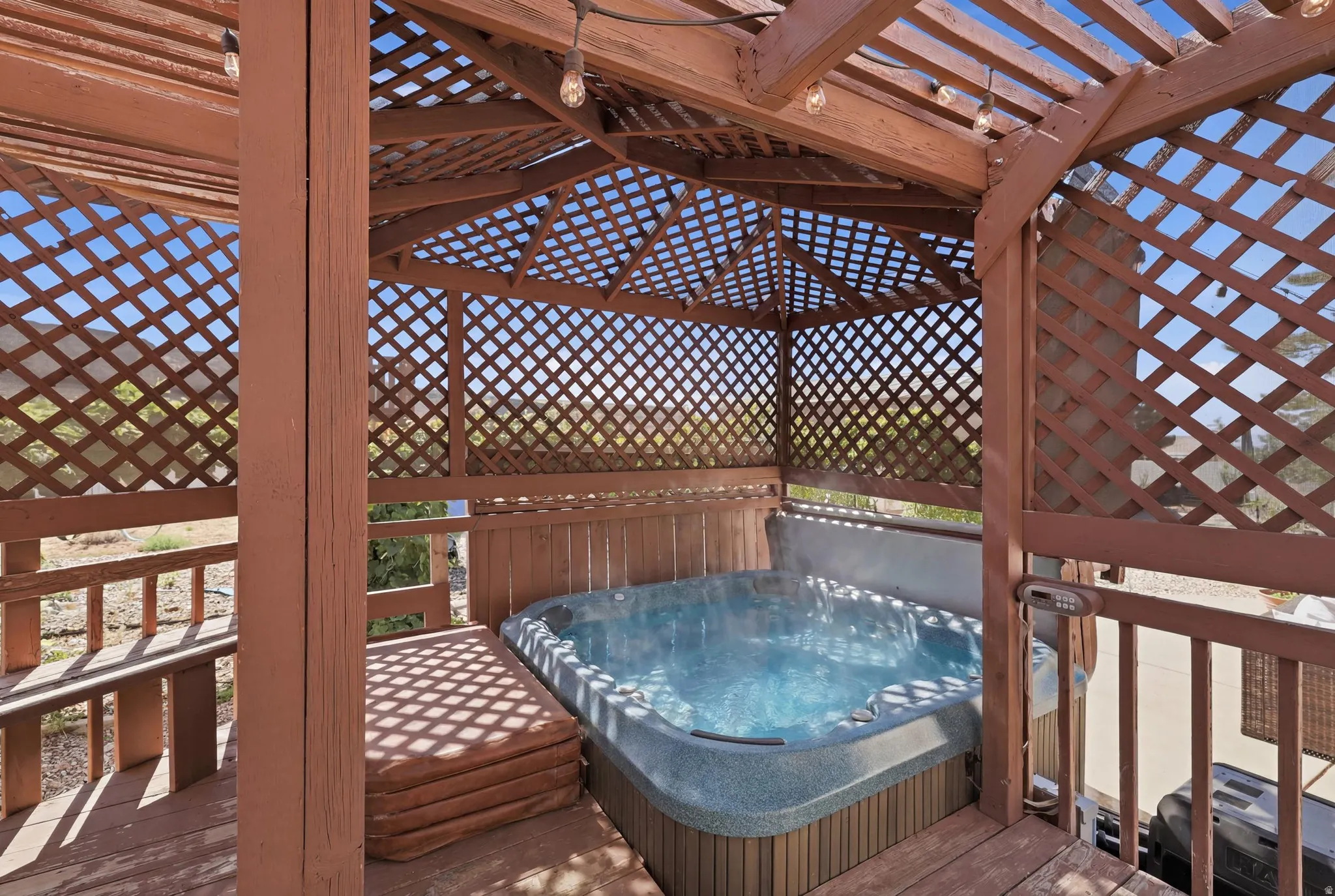 Outdoor spa enclosure featuring a lattice roof and sides, natural wood decking, and string lighting