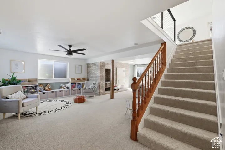 Carpeted staircase with wood newel posts and banister, leading to a lower level featuring recessed lighting, a modern ceiling fan, and a brick accent wall with a built-in fireplace