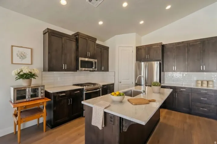 Kitchen featuring dark wood-finish cabinetry, stainless steel appliances, a large center island with an undermount sink, marble-look countertops, and recessed lighting