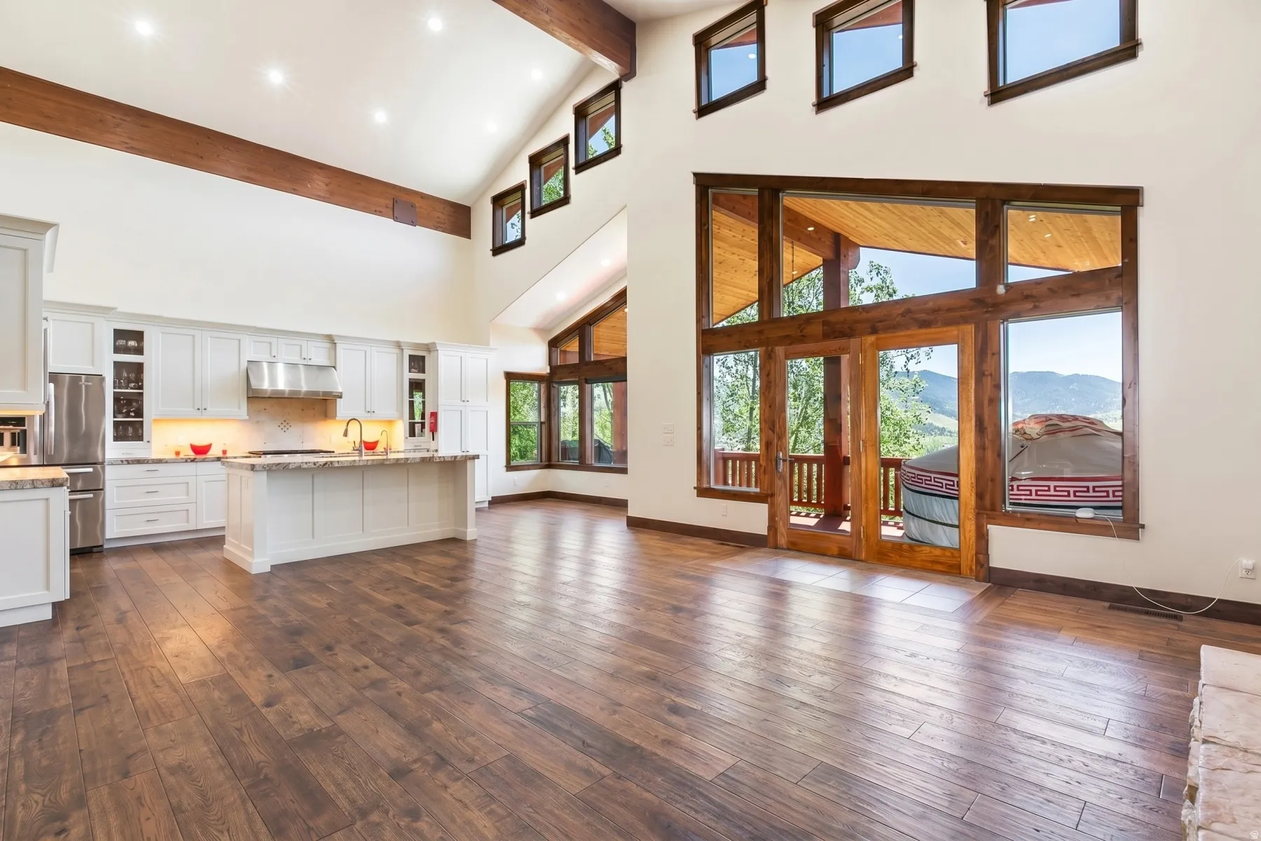 Spacious open-concept living area featuring a vaulted ceiling with exposed wood beams, wood-finish flooring, a contemporary kitchen with white cabinetry and an island, and expansive glass doors opening to outdoor space