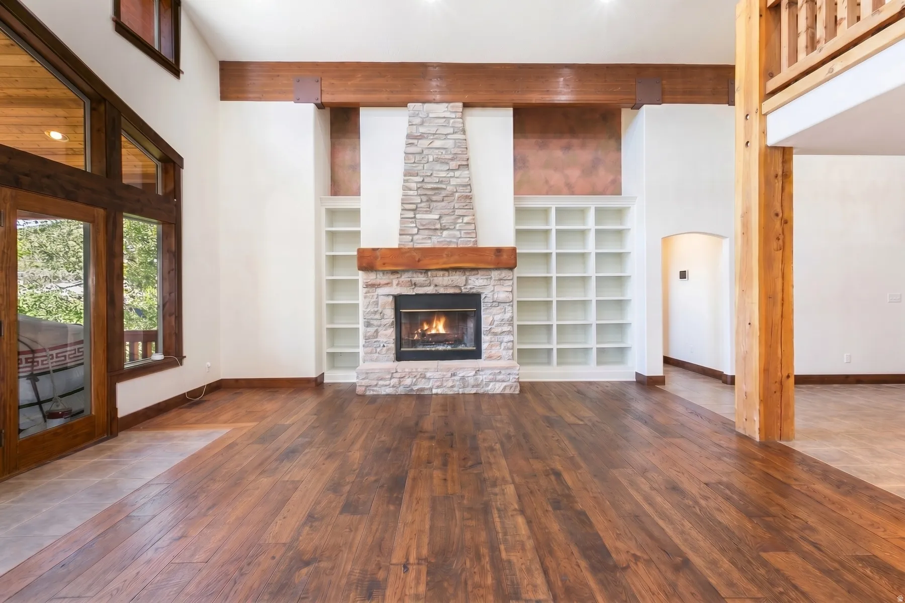 Spacious room featuring wood-finish flooring, a stone-clad fireplace with a wood mantel, built-in shelving, exposed wood beams, and a wood-framed glass door