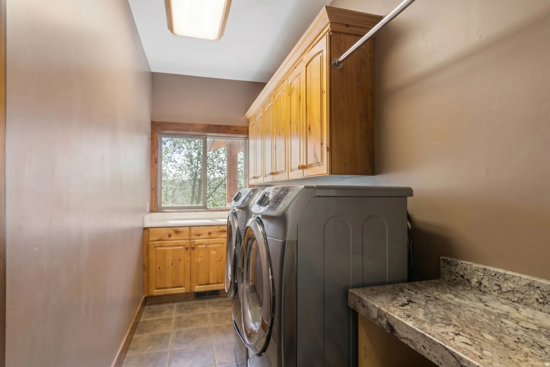 Laundry room featuring wood-finish cabinetry, a utility sink, a window with exterior scenery, a wall-mounted drying rod, and a speckled countertop