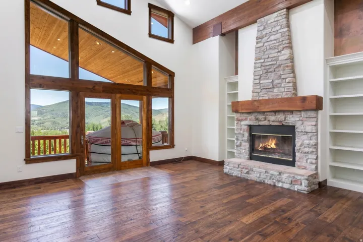 Vaulted living space featuring expansive windows, wood-finish flooring, a stone-clad fireplace with a timber mantel, and built-in shelving