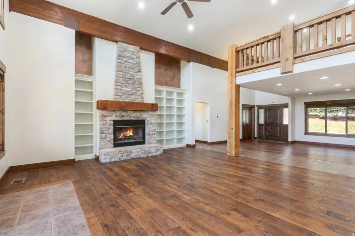 Spacious living area featuring high ceilings with exposed wood beams, a stone fireplace with a wood mantle, extensive built-in shelving, wood-finish flooring, and a loft area with a wood railing