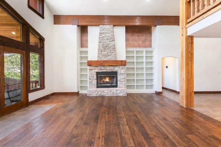 Spacious room featuring wood-finish flooring, a stone-clad fireplace with a wood mantel, built-in shelving, exposed wood beams, and a wood-framed glass door