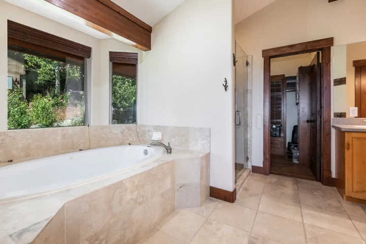 Recessed soaking tub with integrated tiled surround, large windows, wood-finish ceiling beam, glass-enclosed shower, and tile flooring