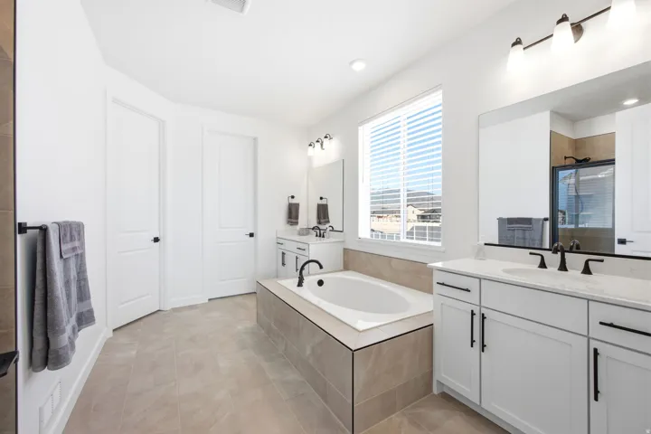 Spacious bathroom featuring a built-in soaking tub with tiled surround, dual vanities with white cabinetry and black hardware, and a large window with blinds