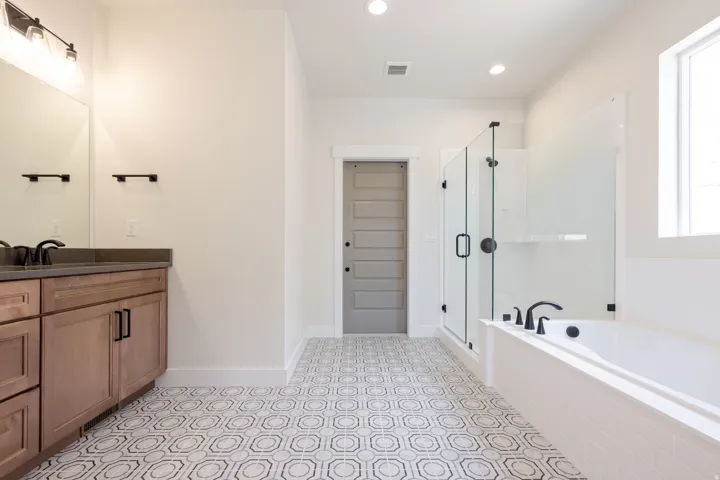 Bathroom featuring a patterned tile floor, a built-in soaking tub with a tiled surround, a glass-enclosed shower, a double vanity with a dark countertop, and matte black fixtures