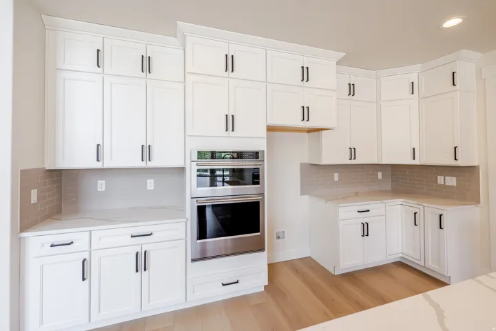 Kitchen featuring white shaker-style cabinetry with matte black hardware, integrated double ovens, light wood-finish flooring, and light-toned subway tile backsplash