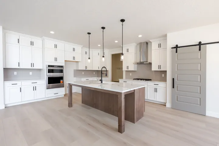 Kitchen featuring a large central island with a stone countertop, white shaker cabinetry with dark hardware, stainless steel built-in appliances, light wood-finish flooring, and a gray barn door