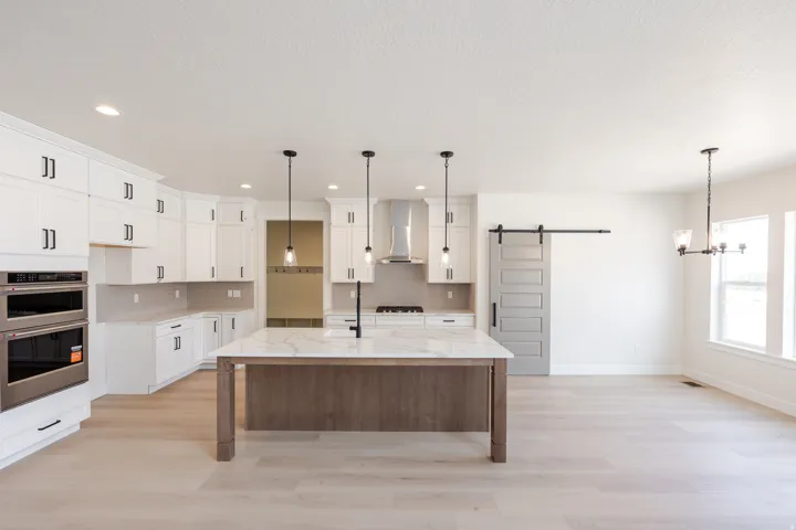 Open-concept kitchen featuring a large central island with a stone countertop, light wood-finish flooring, white cabinetry with black hardware, stainless steel double ovens, and a gray barn door