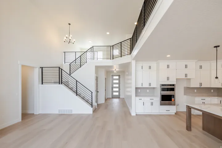 Spacious open-concept interior featuring light wood-finish flooring, a contemporary staircase with black horizontal railings, a two-story vaulted ceiling, recessed lighting, and a modern kitchen with white cabinetry and stainless steel built-in appliances