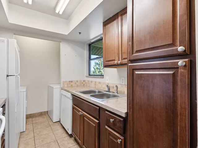 Kitchen area featuring wood-finish cabinetry, a double basin stainless steel sink, light-toned countertop, and a tiled backsplash
