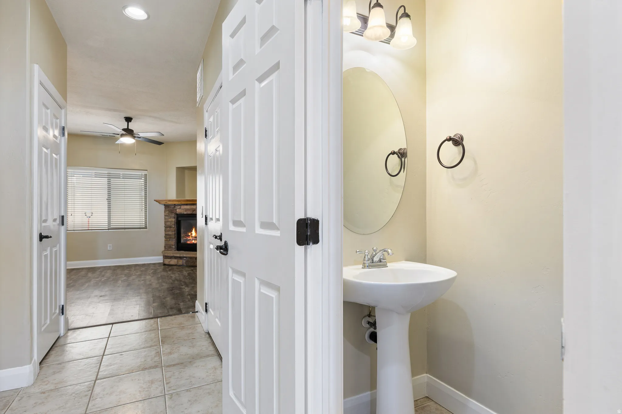 Pedestal sink with an oval mirror and wall-mounted light fixture, complemented by a stone-clad fireplace with a wood-finish mantel and ceiling fan