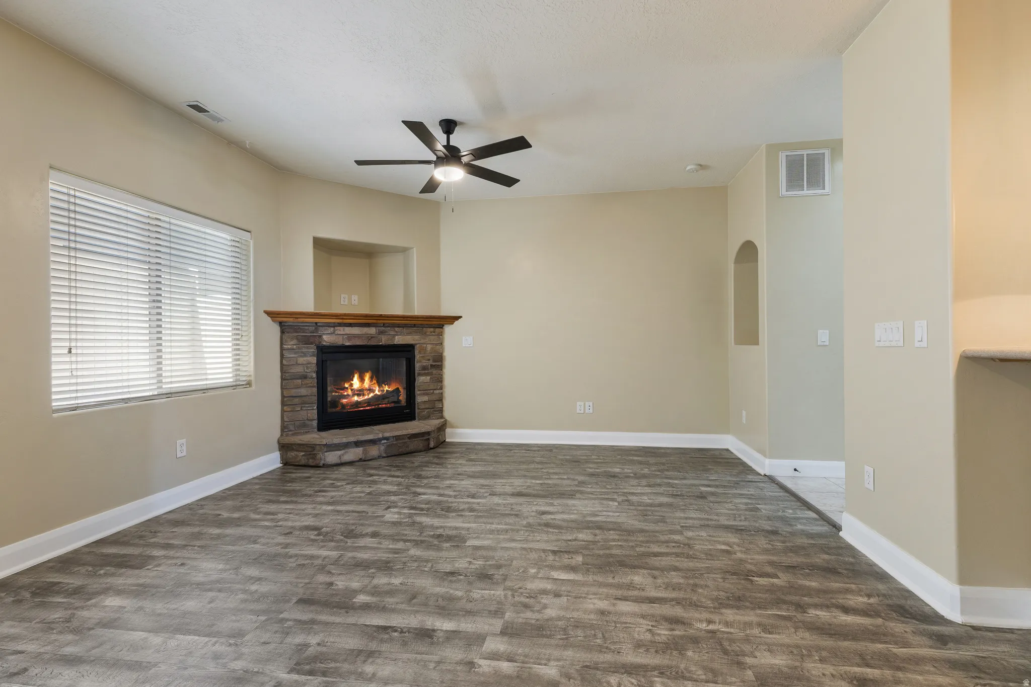 Living area featuring a corner fireplace with a stone surround and wood mantel, wood-finish flooring, a ceiling fan, and a large window with blinds