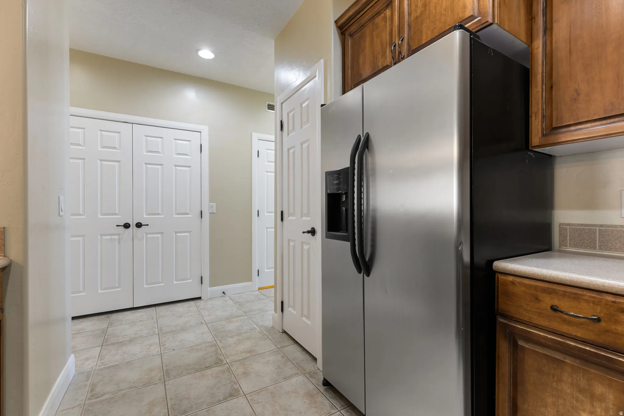 Stainless steel side-by-side refrigerator, warm wood-finish cabinetry, light-toned countertop, tile backsplash, and neutral tile flooring