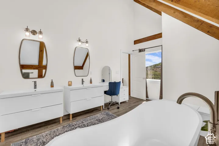 Vaulted ceiling with exposed wood beams, dual floating vanities with integrated sinks, freestanding soaking tub with brushed nickel fixture, wood-finish flooring, and a private balcony access