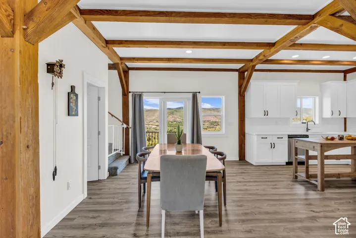 Open-concept living space with exposed wood beams, wood-finish flooring, white walls, and a modern kitchen featuring white cabinetry, a built-in dishwasher, and a stainless steel sink