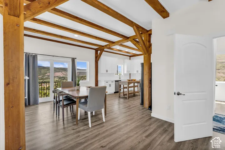 Open-concept living space with exposed wood beams, wood-finish flooring, white cabinetry, and a balcony with mountain scenery