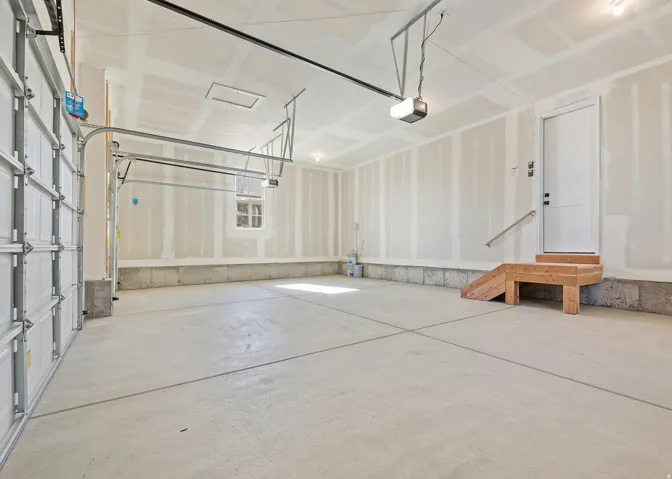 Expansive two-car garage featuring a concrete floor, automatic door opener system, a side access door with wood steps and handrail, a single frosted window, and a utility access panel