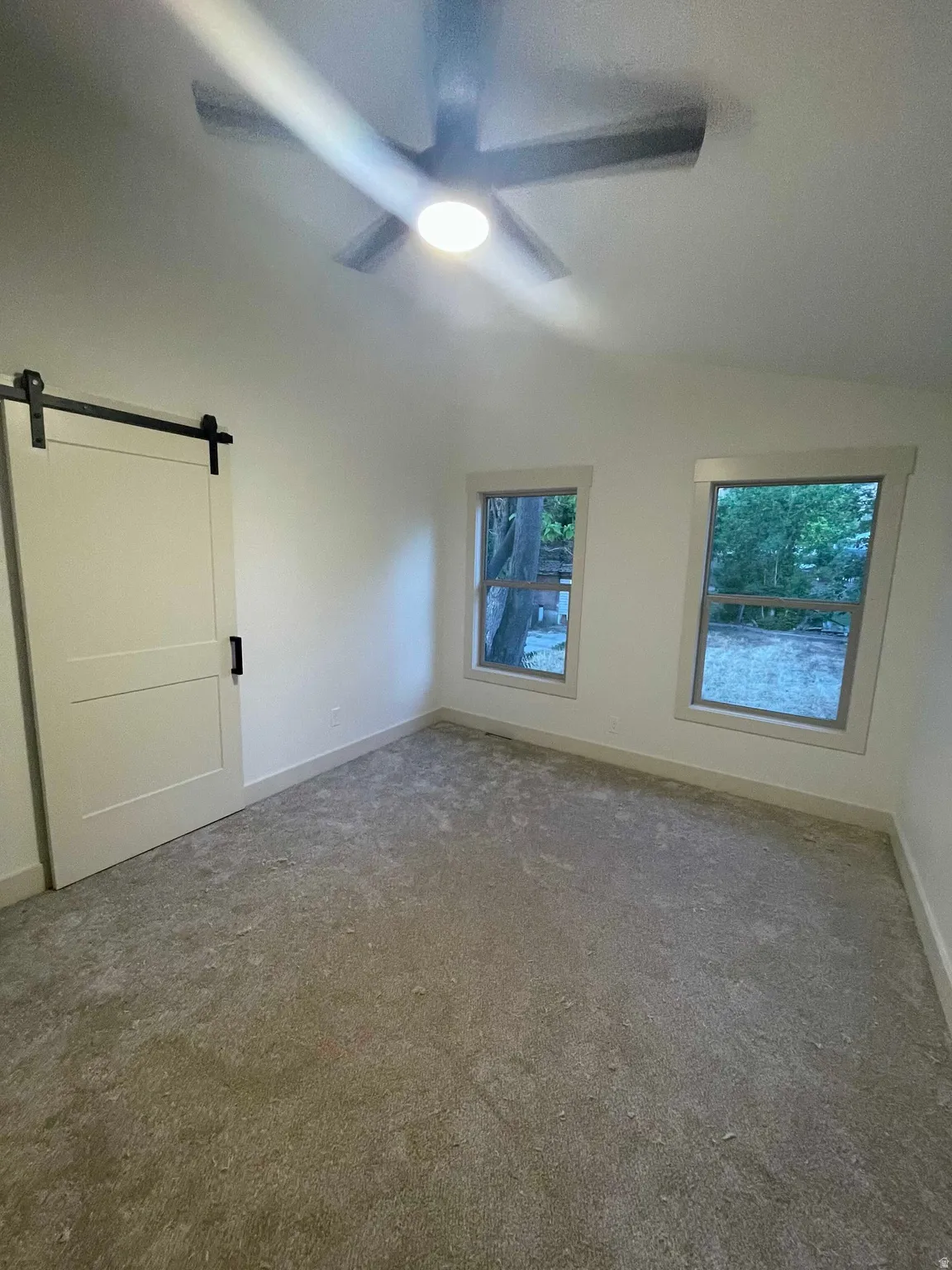 Neutral tone carpeted room featuring a ceiling fan with integrated lighting, two windows, and a modern barn door