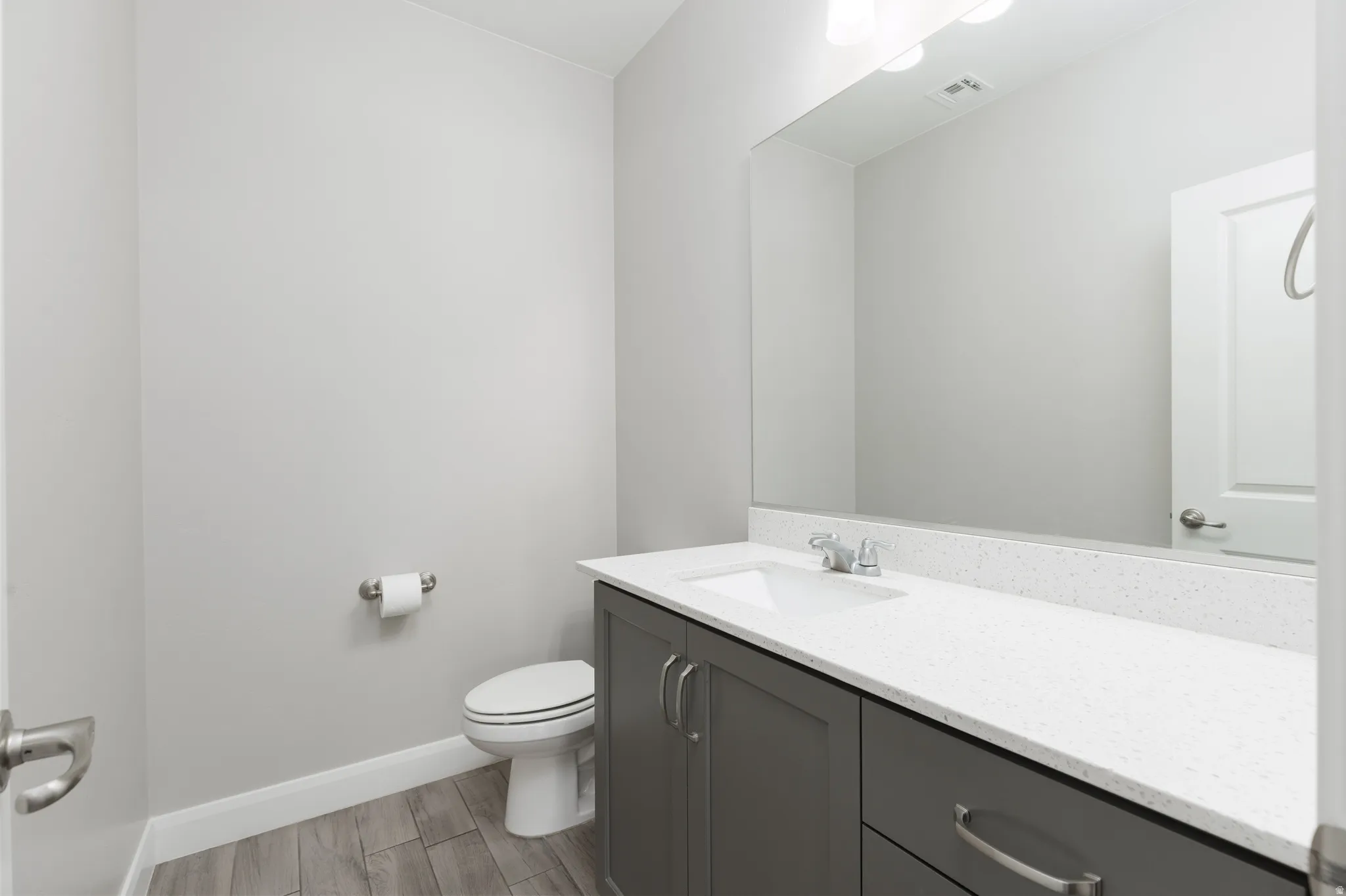 Contemporary bathroom featuring a grey vanity with brushed nickel hardware, a white countertop with integrated sink, a large wall-mounted mirror, and wood-finish flooring