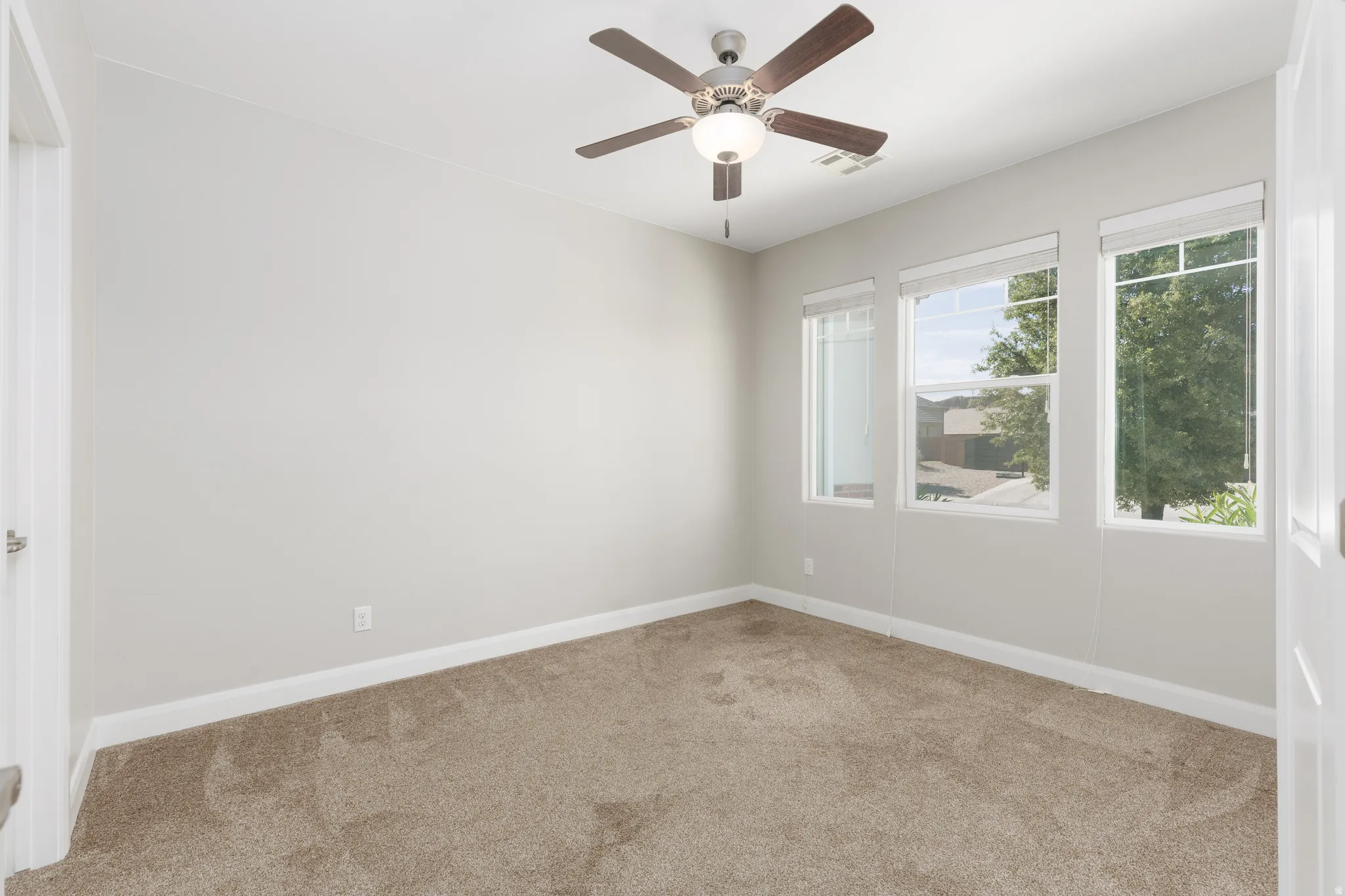 Bright room featuring three large windows, a ceiling fan with integrated lighting, neutral wall paint, and beige carpeting