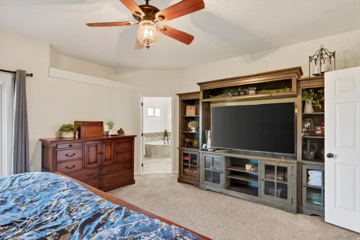 Carpeted room featuring a ceiling fan with integrated lighting, built-in entertainment center, and a doorway opening to a bathroom with a soaking tub