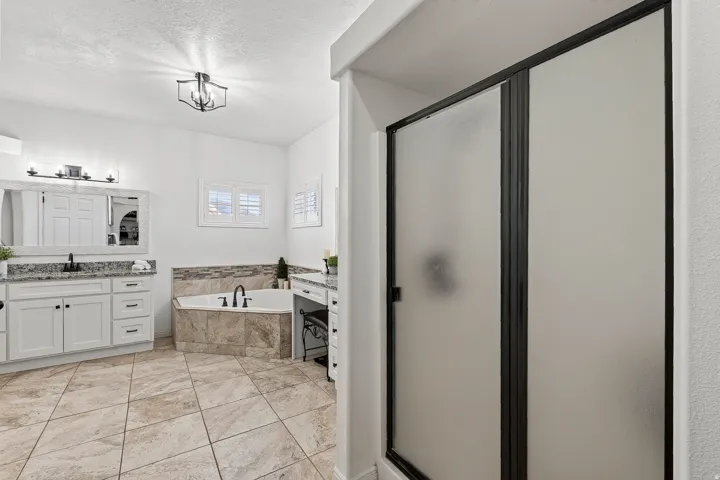Spacious bathroom featuring a corner soaking tub with natural stone tile surround, dual white vanity cabinets with stone countertops, and a separate shower with frosted glass doors