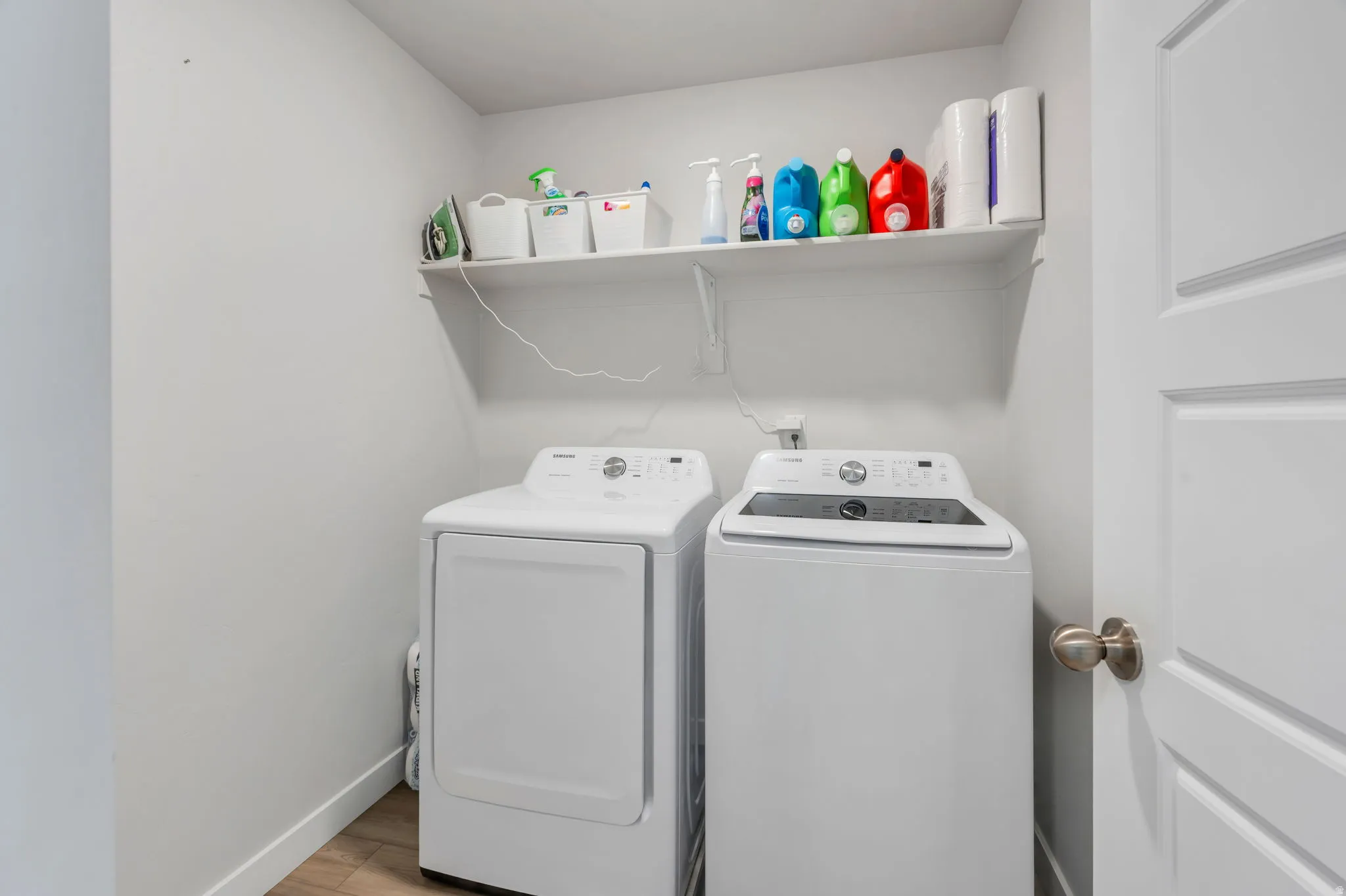 Dedicated laundry space featuring a white wall shelf for storage, wood-finish flooring, and a panel door with a satin nickel doorknob