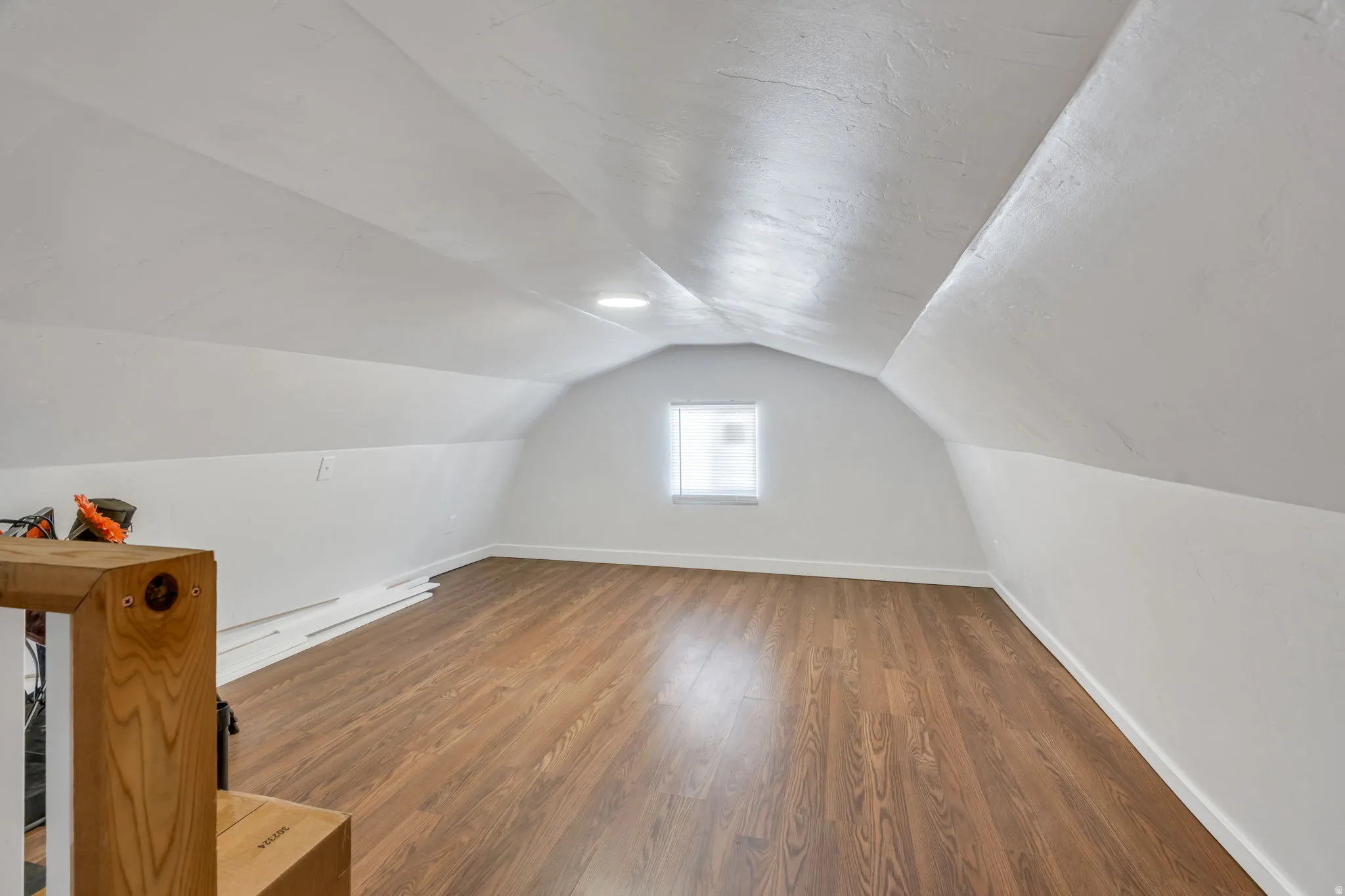 Spacious attic space featuring wood-finish flooring, white walls, and a window with blinds
