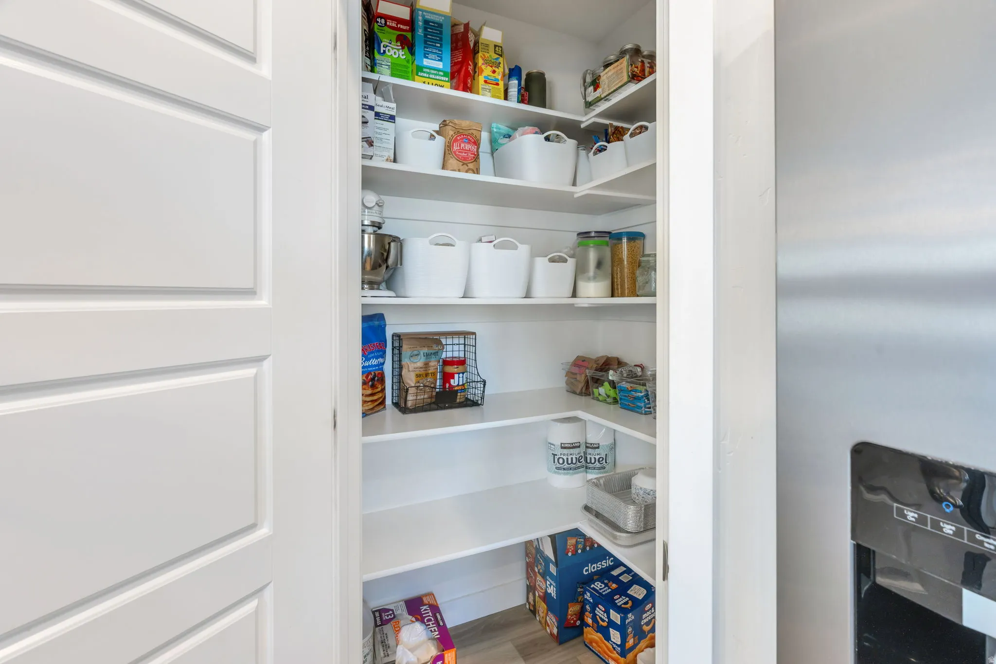 Walk-in pantry with multiple white shelving units, white paneled door, and light wood-finish flooring
