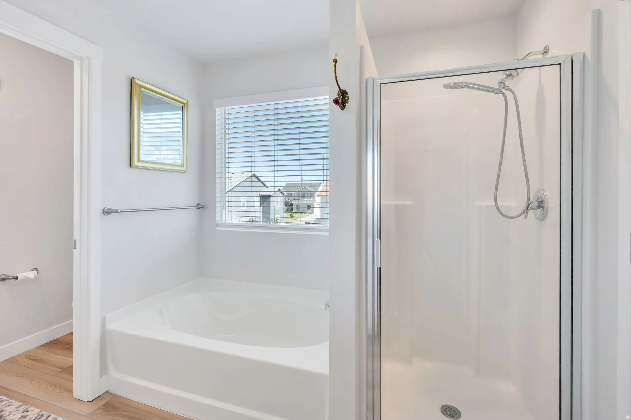 Bathroom featuring a white soaking tub, a separate glass-enclosed shower with a handheld showerhead, light wood-finish flooring, and a window with horizontal blinds