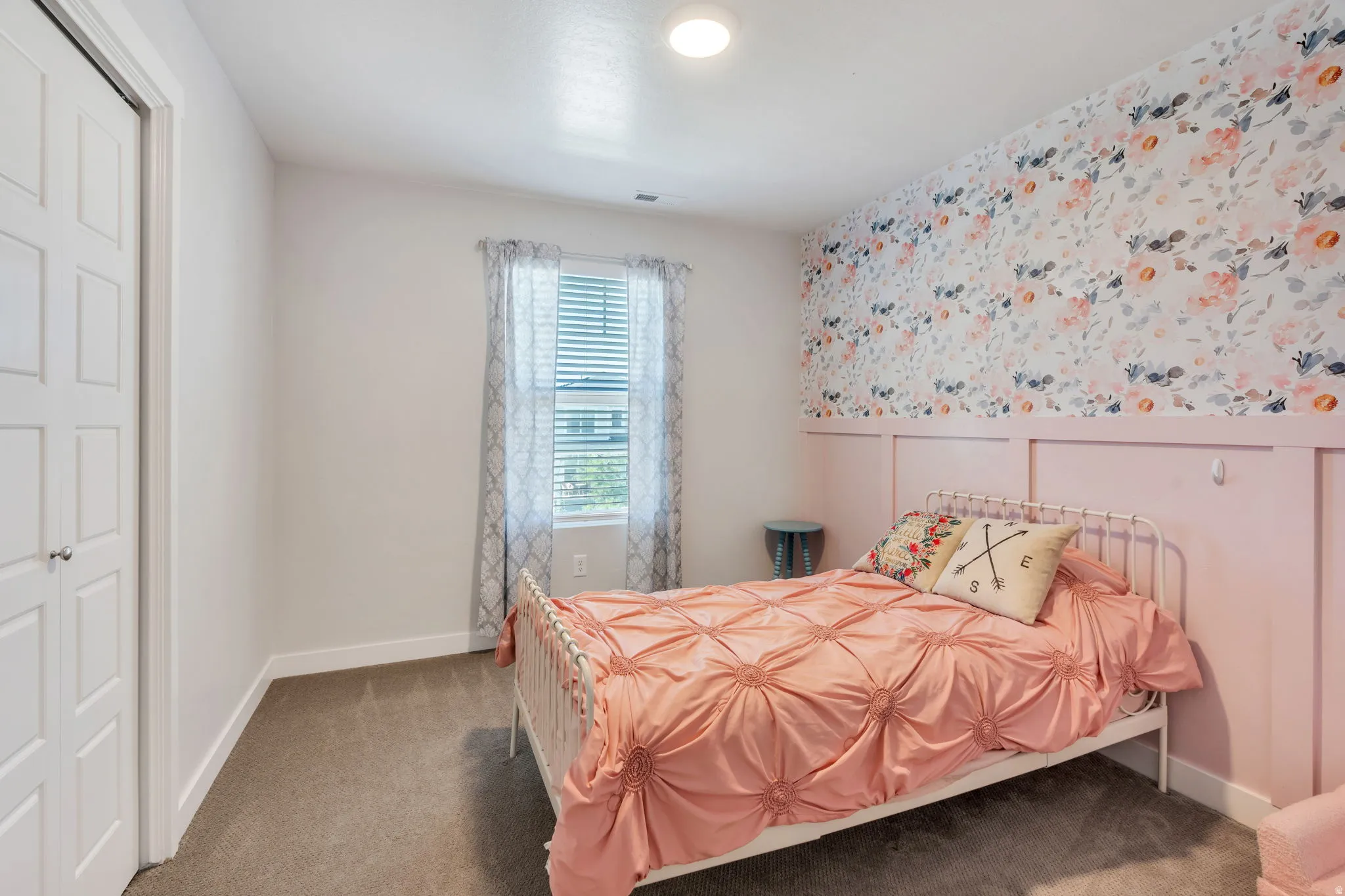 Bedroom featuring a floral accent wall with wainscoting, neutral carpeting, and a double-panel bifold closet door