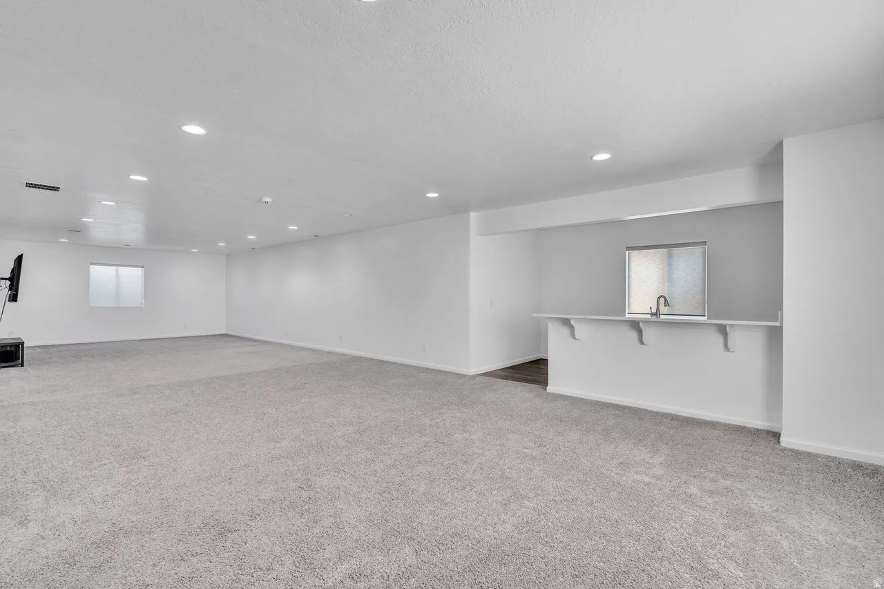 Spacious room featuring light gray carpeting, recessed ceiling lighting, a built-in bar with a sink and window, and white walls