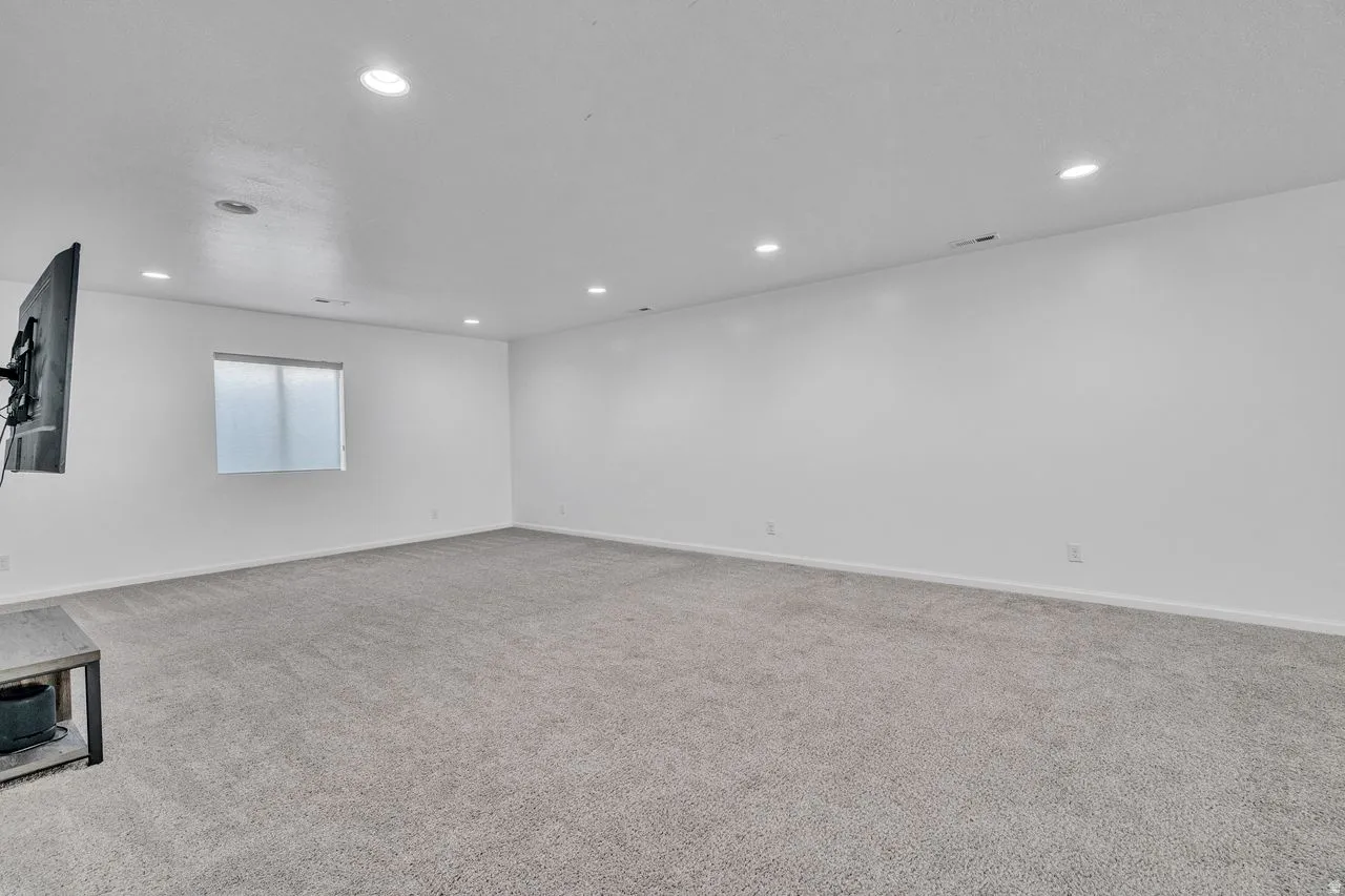 Spacious room featuring light gray carpeting, recessed ceiling lighting, a single window with a privacy screen, and white painted walls