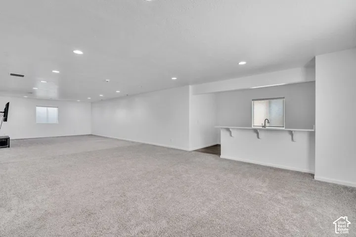 Spacious room featuring light gray carpeting, recessed ceiling lighting, a built-in bar with a sink and window, and white walls