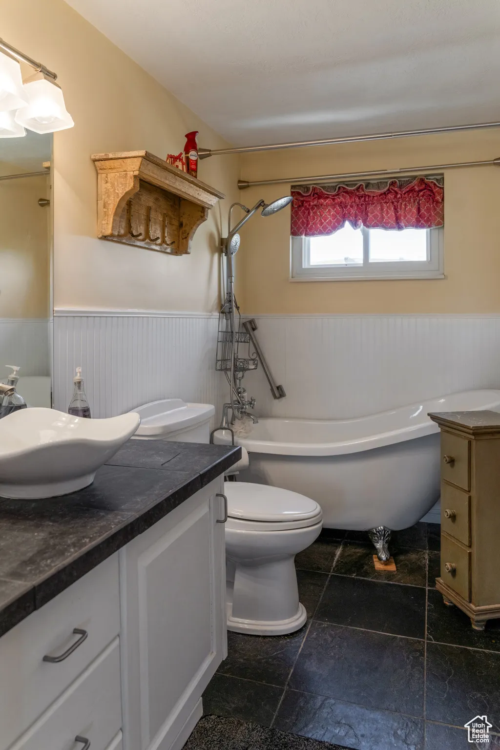 Bathroom featuring a white clawfoot bathtub, black tile flooring, white vanity with a vessel sink, and wainscoting