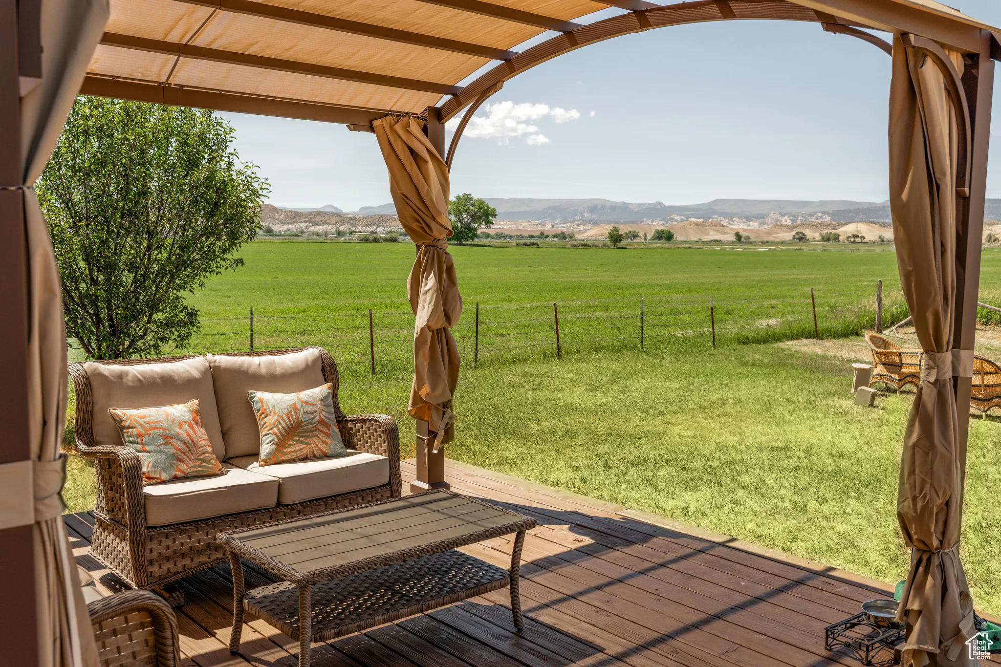 Expansive outdoor deck featuring a shade pergola with fabric canopy and drapes, wood-finish decking, and a wide green field beyond