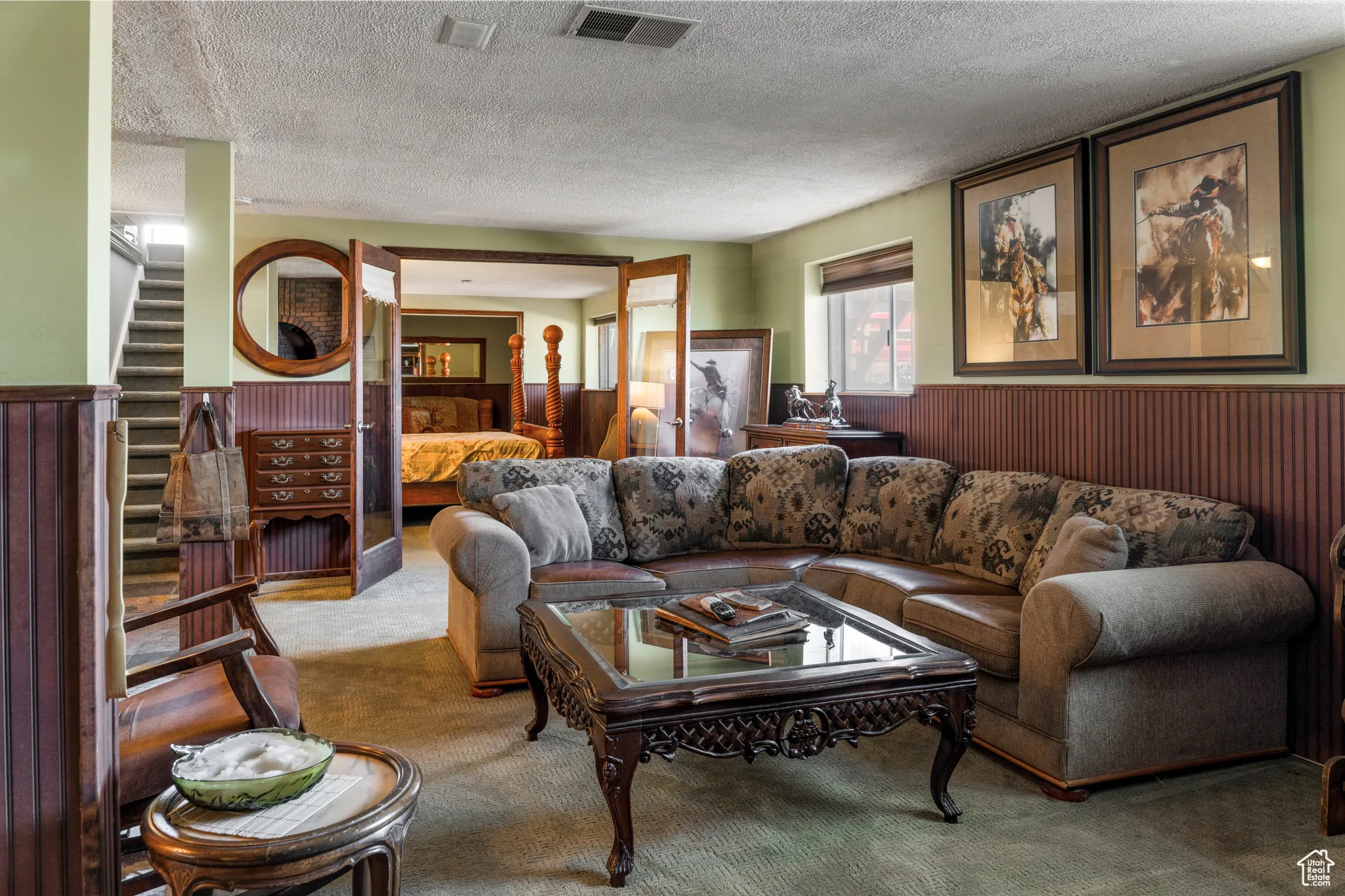 Spacious living area featuring carpet flooring, wood paneling, an exposed staircase, and direct access to an additional room through glass-paneled doors