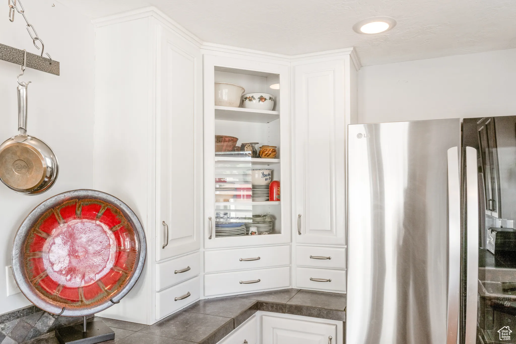 White cabinetry with crown molding, glass-front corner cabinet, under-cabinet drawers, dark countertops, and stainless steel refrigerator