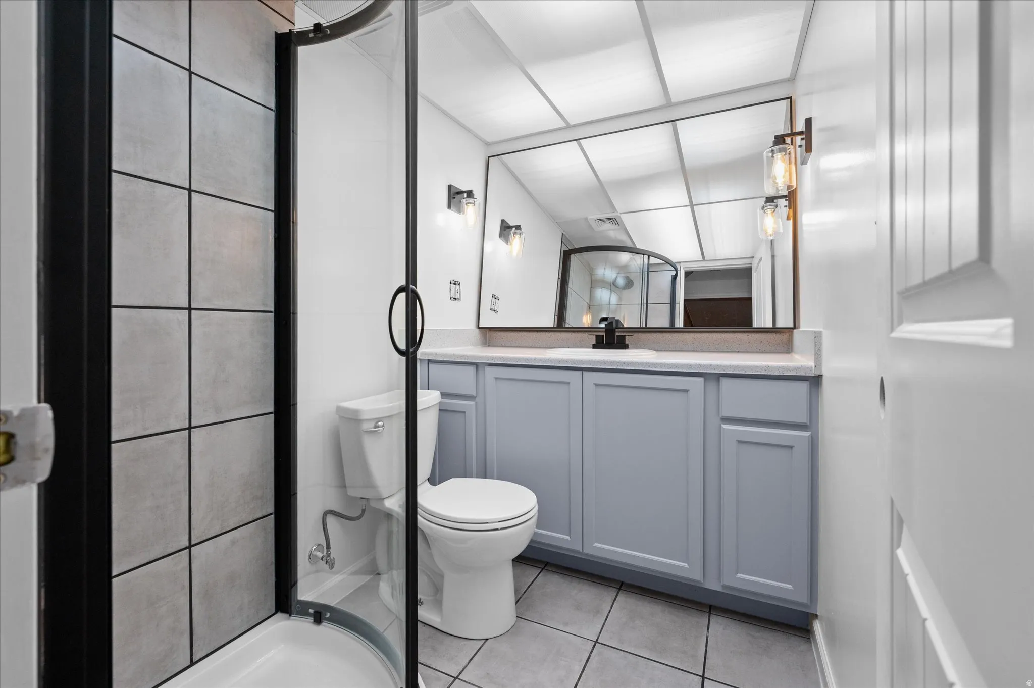 Contemporary bathroom featuring a curved glass shower enclosure with dark framing, a single vanity with gray cabinetry and light countertop, and a large wall-mounted mirror